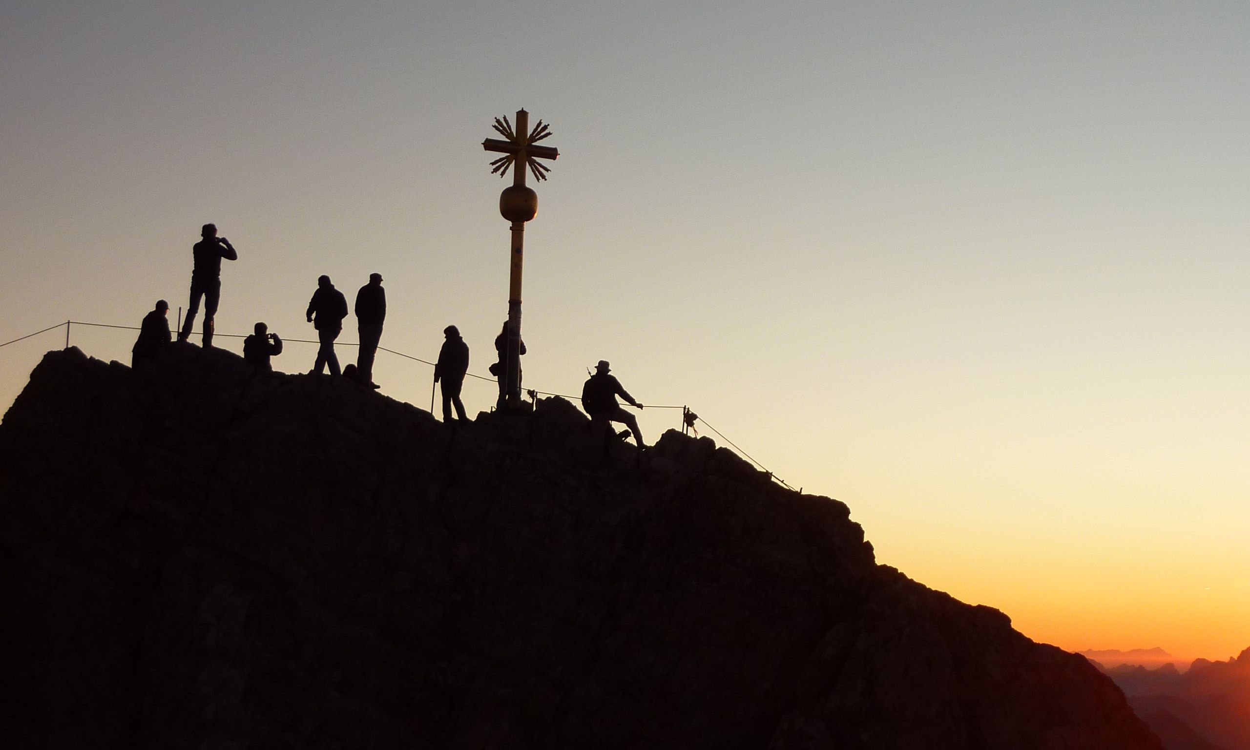 Silhouetted hikers reach a mountain summit at sunrise, with a cross marking the peak.